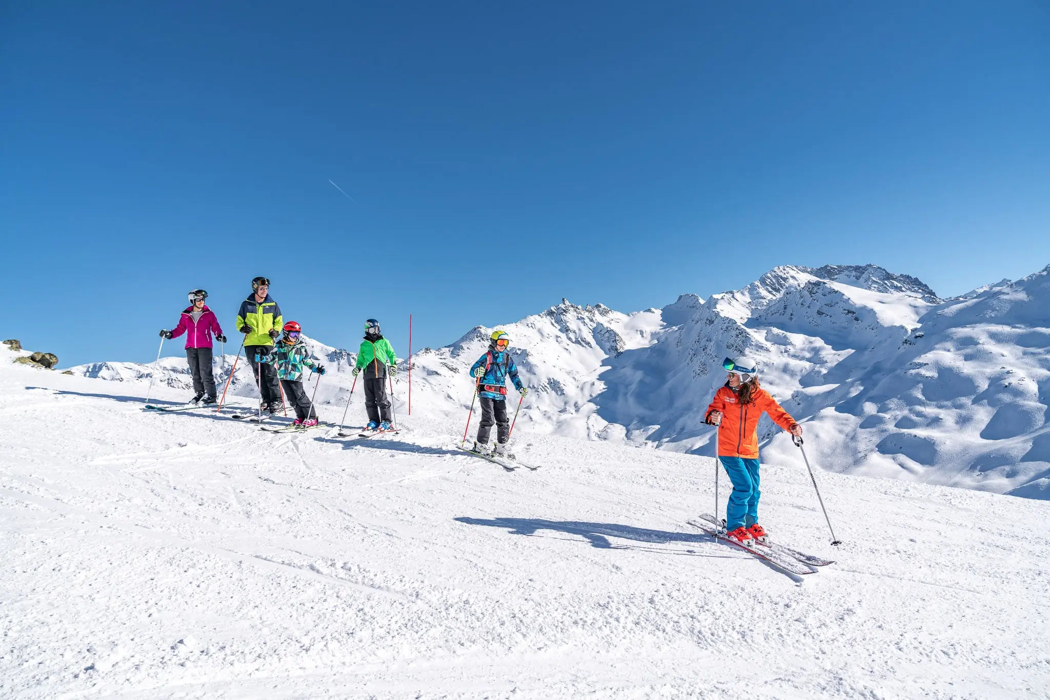 A family with three children is taking a lesson with an Oxygène instructor, dressed in orange and blue. They are standing on a snowy mountain slope under a clear blue sky.